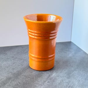 May include: A vibrant orange ceramic vase with a glossy finish, featuring a wide, flared rim and two distinct horizontal ridges encircling the upper and lower sections of the cylindrical body, creating a simple, functional vessel design.