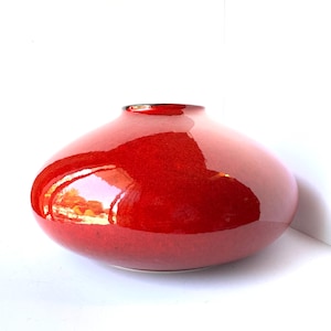 May include: A low, wide-bodied vase with a smooth, glossy finish in a vibrant, deep red color. The vessel features a rounded, bulbous shape that tapers toward a small, circular opening at the top, reflecting light across its polished surface.