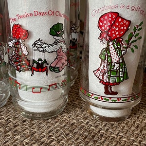 May include: Clear glass tumblers with Christmas-themed illustrations. One tumbler features a child with a puppet, another shows a girl with holly. Text includes "The Twelve Days Of Christmas" and "Christmas is a gift of joy."