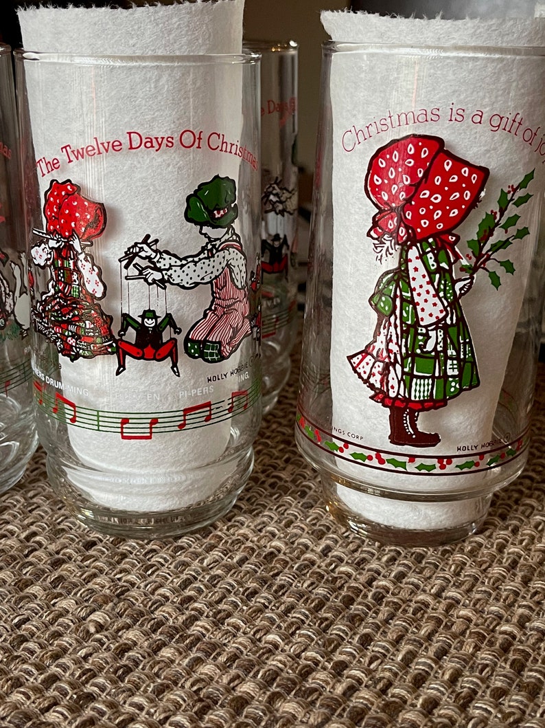 May include: Clear glass tumblers with Christmas-themed illustrations. One tumbler features a child with a puppet, another shows a girl with holly. Text includes "The Twelve Days Of Christmas" and "Christmas is a gift of joy."