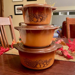 May include: A stack of three brown casserole dishes with lids. The dishes have a gradient color and a fruit design. The lids and handles are a darker shade of brown. The dishes are stacked on a wooden table.