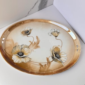 May include: Oval ceramic serving tray with a gold rim and handles. The tray features hand-painted poppy flowers in shades of cream, brown, and black. The flowers are arranged on a cream-colored background, with brown leaves and stems.
