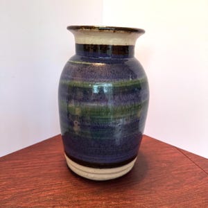 May include: A handcrafted ceramic vase with a rounded body and a slightly flared rim. The vase features horizontal bands of color, including blue, green, and cream, with a dark brown rim and base. The vase is approximately 8 inches tall.