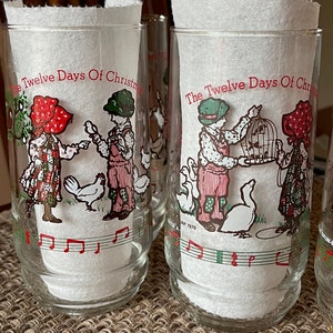 May include: Set of clear glass tumblers featuring illustrations from "The Twelve Days of Christmas." Each glass has a different scene with children, animals, and musical notes in red, green, and white. The glasses are filled with white paper.