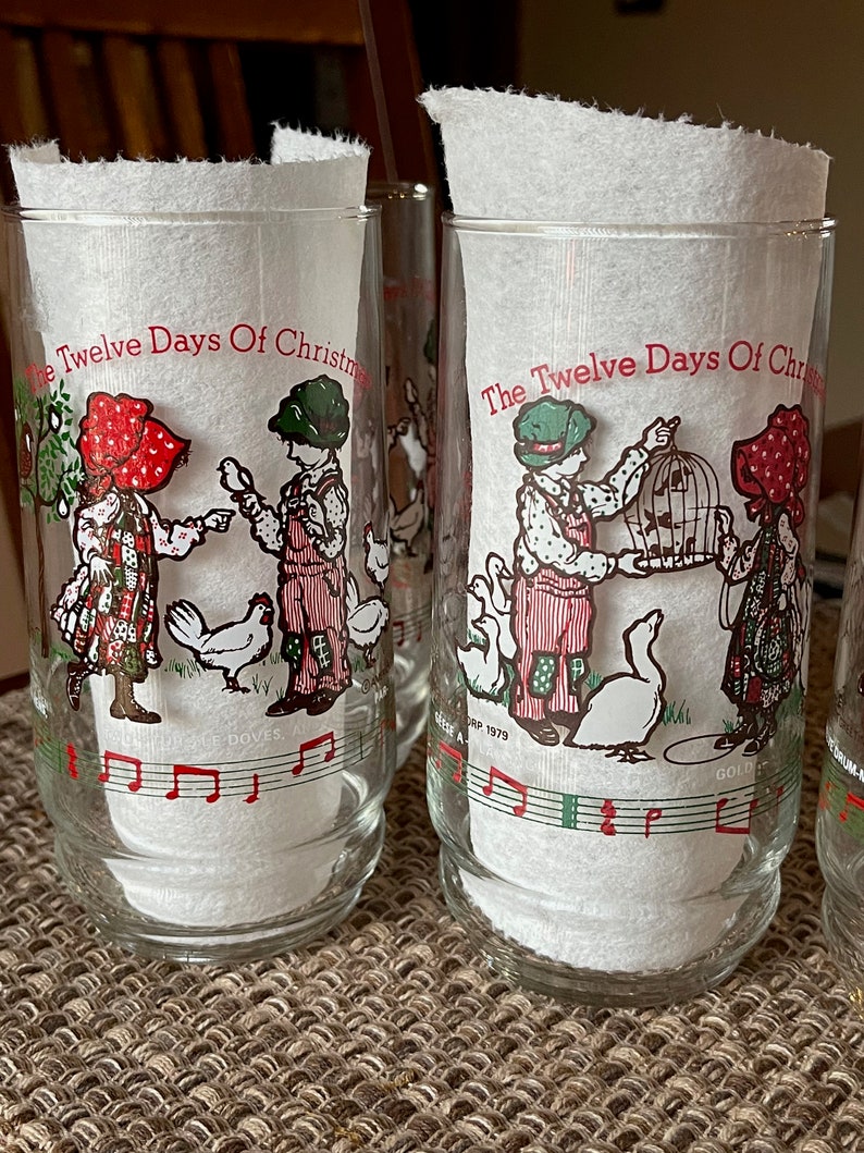 May include: Set of clear glass tumblers featuring illustrations from "The Twelve Days of Christmas." Each glass has a different scene with children, animals, and musical notes in red, green, and white. The glasses are filled with white paper.