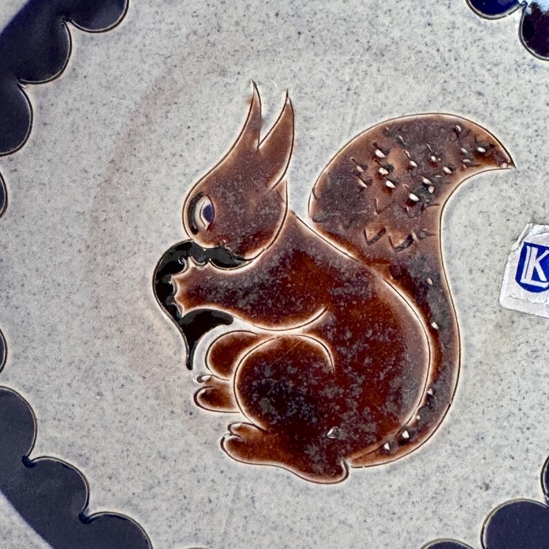May include: A ceramic plate with a brown squirrel design. The squirrel is holding a black object and has a fluffy tail with white speckles. The plate has a blue and white border.