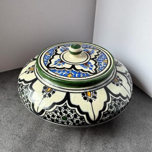 May include: A round ceramic lidded jar featuring a cream-colored base with a crackled glaze finish. The surface is decorated with intricate black, blue, green, and yellow floral and geometric patterns. It includes a matching lid with a knob.