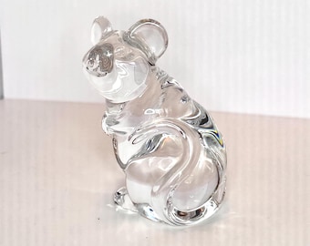 May include: A clear glass figurine of a mouse in a seated position. The mouse has large ears and a rounded body. The figurine is made of transparent glass, allowing light to pass through, and is displayed on a white surface.