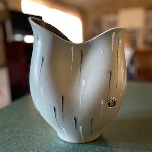 May include: A light gray ceramic vase with a flared rim and a dark interior. The vase features vertical streaks of black and white paint. A small sticker with a crown and the letters EIR is visible on the side. The vase is on a green surface.