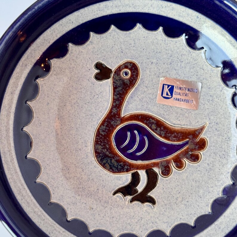 May include: Ceramic plate with a blue rim and a light gray center. The plate features a brown and purple bird design. The plate has a scalloped edge and a sticker that reads "FEINSTEINZEUG QUALITAT HANDARBEIT."
