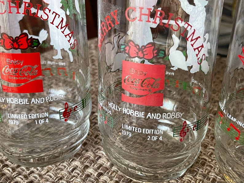 May include: Clear glass Coca-Cola Christmas glasses. Each glass features a red Coca-Cola logo, the words "Merry Christmas", and "Holly Hobbie and Robby". Limited edition, 1979.