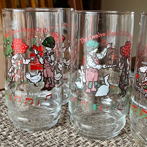 May include: Set of clear glass tumblers featuring illustrations from "The Twelve Days of Christmas" carol. Each glass has a different scene with children, birds, and musical notes in red, green, and white. The text on the glasses includes song lyrics.