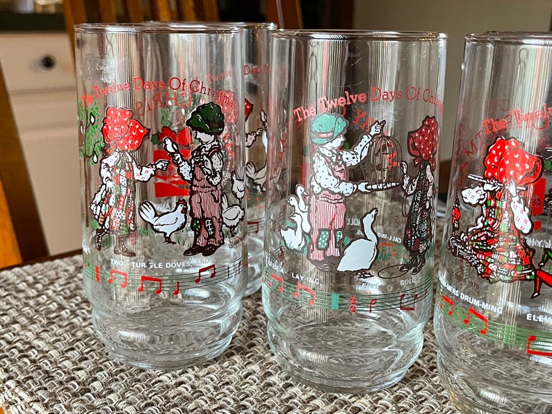 May include: Set of clear glass tumblers featuring illustrations from "The Twelve Days of Christmas" carol. Each glass has a different scene with children, birds, and musical notes in red, green, and white. The text on the glasses includes song lyrics.