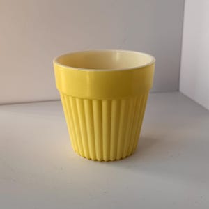 May include: A small, yellow, ribbed planter with a cream-colored interior. The planter has a cylindrical shape with vertical ridges along the exterior. The rim is smooth and the base is flat. This container is suitable for small plants or succulents.