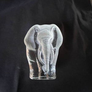 May include: A clear glass elephant figurine with detailed trunk, ears, and textured body. The sculpture is standing upright and appears to be made of a single piece of glass. The elephant is centered against a dark background.