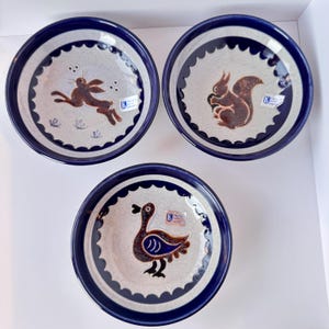 May include: Three ceramic plates with a blue rim and animal illustrations. One plate features a brown rabbit, another a squirrel, and the third a duck. The plates have a light gray center with a scalloped edge.