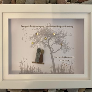 Op de afbeelding: Een ingelijst kunstwerk met een witte rand. De afbeelding toont een boom met gouden accenten en twee kiezelstenen figuren op een schommel. Tekst luidt "Congratulations on your Golden Wedding Anniversary" en namen met een datum.