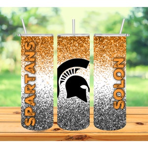 May include: A set of three stainless steel tumblers with lids and straws. The tumblers feature a glitter design in orange, white, and black. The word "SPARTANS" is printed vertically on one tumbler, and "SOLON" on another. A black Spartan helmet logo is on the center tumbler.