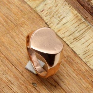 Op de afbeelding: Een roségouden ring met een hartvormige voorkant. De ring is gemaakt van glanzend metaal en ligt op een houten oppervlak. De hartvorm is glad en gepolijst, waardoor het licht reflecteert. De band van de ring is dik en solide.