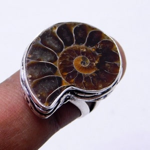 May include: A silver ring with a large, brown ammonite fossil set in the center. The ammonite has a spiral pattern with a dark brown center and lighter brown outer edges.