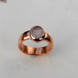 Peut inclure: Une bague en cuivre avec une pierre précieuse ovale rose clair. La bague a un large anneau et un design simple et élégant. La pierre précieuse est sertie dans un sertissage à lunette, et le cuivre a une finition polie.