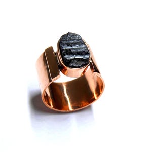 Pode incluir: Um anel cor de cobre com uma pedra preciosa de turmalina negra bruta. O anel tem uma banda larga com um acento retangular de um lado, e a pedra é engastada em uma moldura oval. O anel é exibido em um fundo branco.