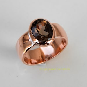May include: A rose gold ring with a large, oval-shaped, smoky brown gemstone. The ring has a wide band and a polished finish. The gemstone is set in a bezel setting. The ring's design highlights the gemstone's colour and shape.