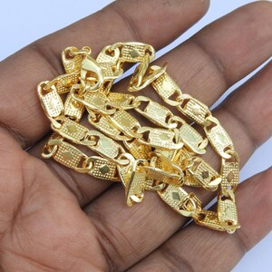 May include: A gold-colored chain necklace with a textured, rectangular link design. The necklace is made of a shiny, metallic material and is displayed on a hand. The links have a repeating pattern of small, square details.