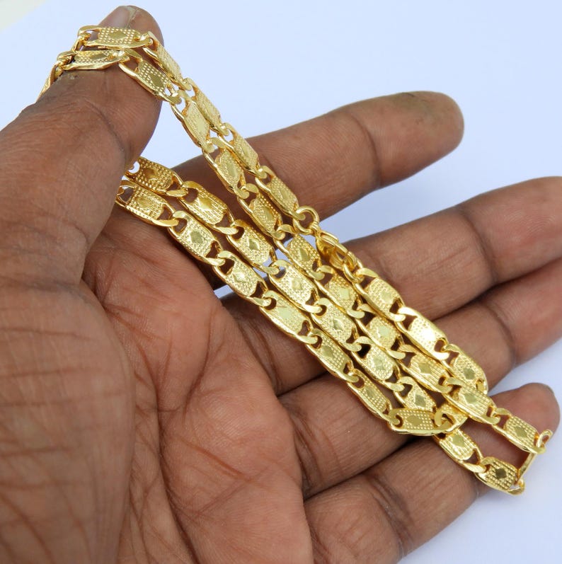 May include: A gold-colored chain necklace with a repeating rectangular link design. The links have a textured pattern and are connected by smaller, curved links. The necklace is displayed on a light background.