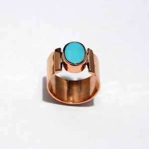 Peut inclure: Une bague de couleur cuivre avec une pierre ovale turquoise vif. La bague a un large anneau avec des accents rectangulaires verticaux de chaque côté de la pierre. La bague est sur fond blanc.