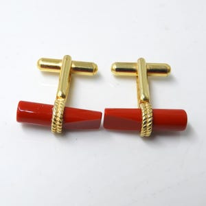 Puede incluir: Un par de gemelos dorados con un centro cilíndrico rojo brillante. La parte dorada tiene un diseño en forma de T. La parte roja está conectada al oro con un diseño retorcido en forma de cuerda.