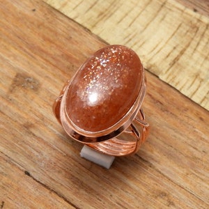 May include: A copper ring with a large oval-shaped sunstone gemstone. The ring has a delicate, twisted band.