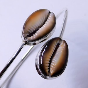 Könnte beinhalten: Ein silbernes Armreif-Armband mit zwei Kaurischnecken. Die Muscheln sind braun und weiß mit einem glänzenden Finish.