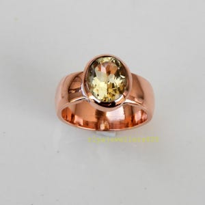 May include: A rose gold ring featuring a large, oval-cut, yellow gemstone. The ring has a wide band and a polished finish. The gemstone is set in a bezel setting, and the ring is displayed on a white surface.