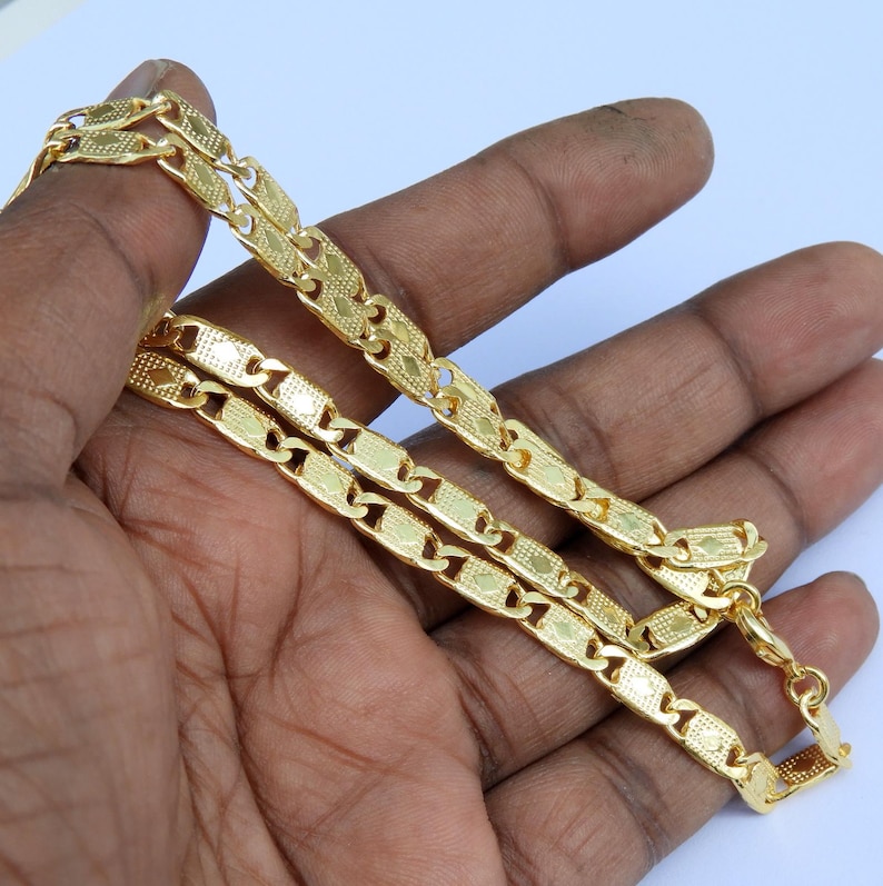 May include: A gold-colored chain necklace with a repeating rectangular link design. The necklace is held in a hand, showcasing its intricate pattern and shiny finish. The chain is approximately 20 inches long.