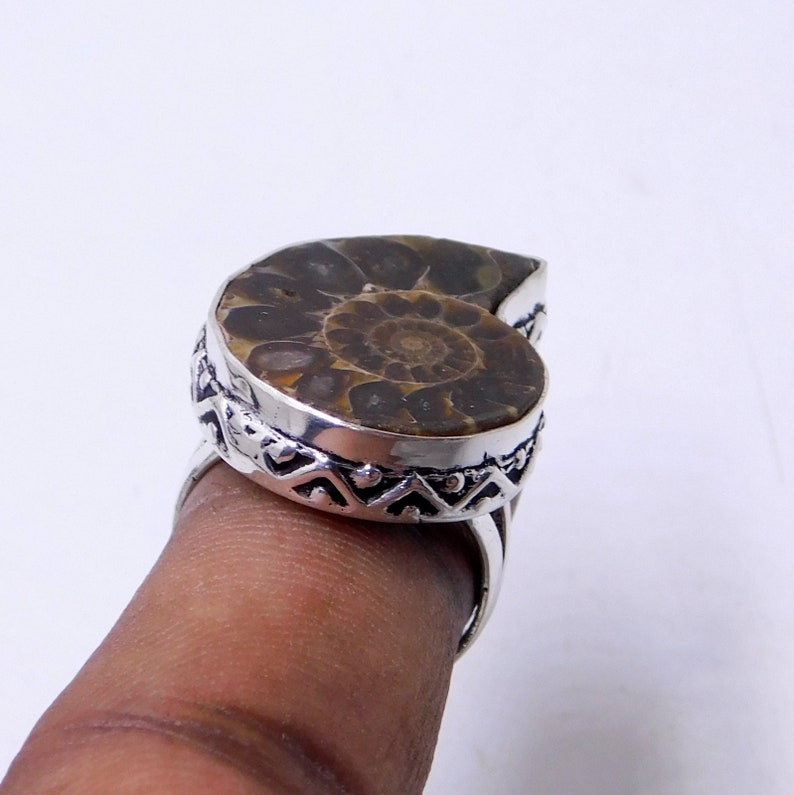 May include: A silver ring with a brown ammonite fossil set in the center. The ring has a decorative band with a geometric pattern.