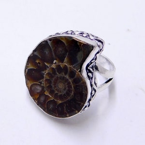 May include: A silver ring with a brown ammonite fossil set in the center. The ring has a detailed, textured band.
