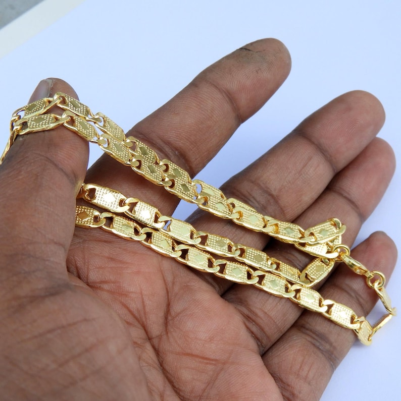 May include: A gold-colored chain necklace with a rectangular link design. The chain is held in a hand against a white background. The links have a textured surface and are connected by smaller, decorative links.