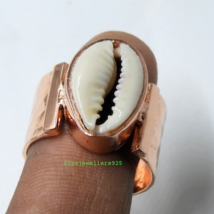 May include: A copper-colored ring featuring a cream-colored seashell centerpiece. The ring has a wide band with a rectangular design on each side of the shell. The seashell is oval-shaped and has a textured surface.