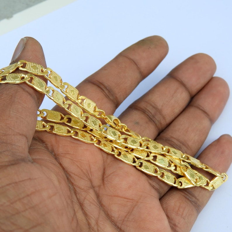 May include: A gold-colored chain necklace with a repeating rectangular link design. The chain is held in a hand against a white background. The links have a textured pattern.