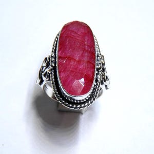 May include: A silver ring with a large, oval-shaped ruby gemstone. The ruby is a bright red hue and is set in an intricate silver band with a twisted rope pattern around the stone. The ring is presented on a white background.