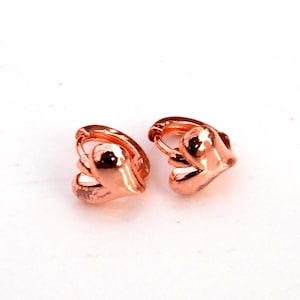 Puede incluir: Dos pendientes de aro en forma de corazón en oro rosa. Los pendientes son pequeños y tienen un acabado pulido. El diseño del corazón es detallado y tridimensional, con una superficie lisa y reflectante. Los pendientes están sobre un fondo blanco.