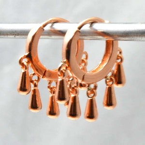 Op de afbeelding: Een paar roségouden oorringen met bungelende druppelvormige bedels. De oorbellen zijn gemaakt van glanzend metaal en hebben een eenvoudig, elegant ontwerp. De druppelvormige bedels voegen beweging en visuele interesse toe.