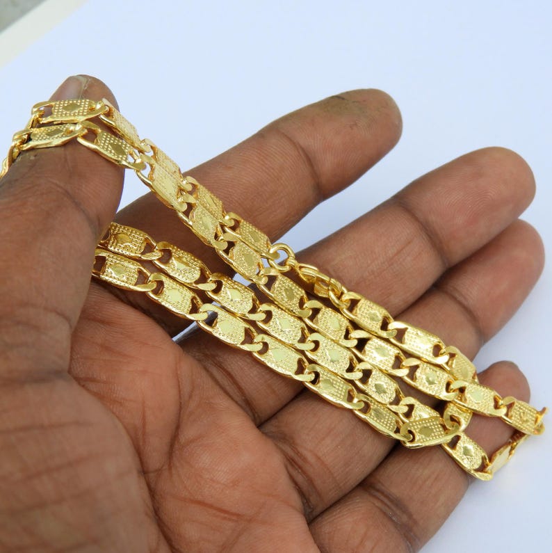 May include: A gold-colored chain necklace with a repeating rectangular link design. The necklace is held in a person's hand, showcasing its intricate pattern and shiny finish. The chain appears to be made of metal.