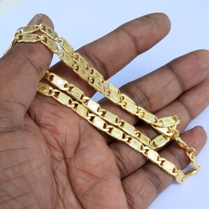 May include: A gold-colored chain necklace with a repeating rectangular link design. The links have a textured surface and are connected by smaller, rounded links. The necklace is displayed against a light background.