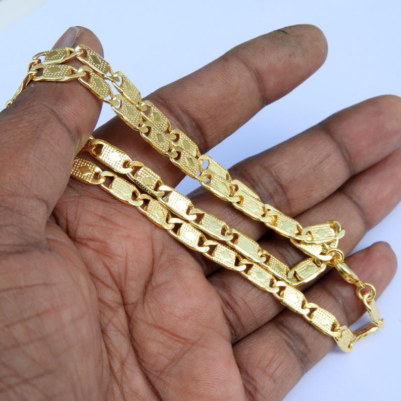 May include: A gold-colored chain necklace with a repeating rectangular link design. The links have a textured surface and are connected by smaller, rounded links. The necklace is displayed against a light background.