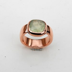 以下が含まれることがあります： 滑らかで丸みを帯びた仕上げの磨かれた銅製のバンドを備えたリング。リングの中央には、淡い半透明の緑色の色合いをした、単一の正方形のファセットカットされた宝石がセットされており、シンプルな銅製のベゼルセッティングでしっかりと固定されています。