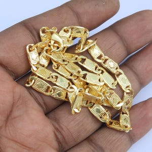 May include: A gold-colored chain necklace with a textured, rectangular link design. The necklace is displayed on a hand, showcasing the intricate details of the jewelry. The clasp is visible, and the chain has a bright, reflective finish.
