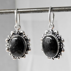 Op de afbeelding: Een paar zilverkleurige oorbellen met ovale, zwarte edelstenen. De edelstenen zijn gezet in een decoratief montuur met een geschulpte rand. De oorbellen hangen aan een eenvoudige oorring. De sieraden worden tegen een neutrale achtergrond getoond.
