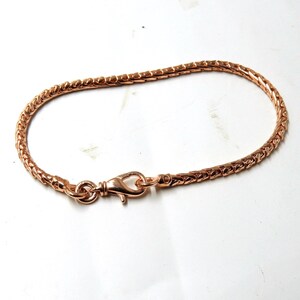May include: Rose gold bracelet with a woven chain design. The bracelet has a lobster clasp closure. The jewellery is displayed on a white surface. The bracelet is a delicate accessory.